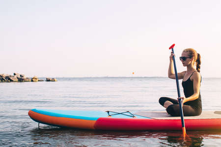 Close-up of a girl floating on a sup board. The concept of water sports, relaxation and self-immersion. Lonely woman alone on a board against a background of water and a pier.の写真素材