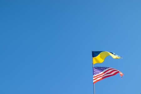 Ukrainian and American flag on a blue sky background. Friendship of peoples and diplomatic mission. Yellow-blue and stars-striped fabric on a flagpole.の写真素材