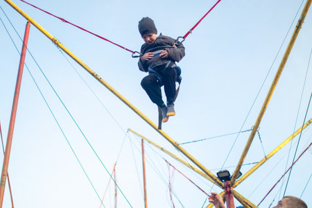 The boy is jumping on a bungee trampoline. A child with insurance and stretchable rubber bands hangs against the sky. The concept of happy childhood and games in the amusement park.の写真素材