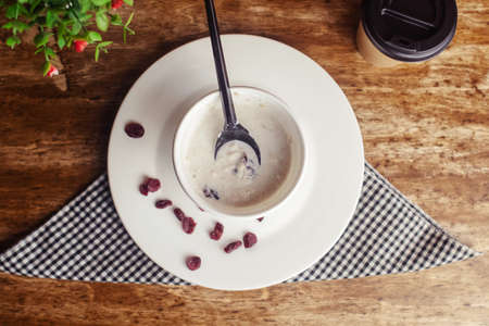 Close-up of oatmeal in a paper cup. Takeaway food. Healthy breakfast with raisins, dried apricots and milk. Wooden background. Nearby is coffee to goの写真素材