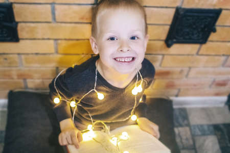 Christmas background. The boy is sitting near the fireplace with a book. Child with yellow lights. Waiting for the new year. Christmas illumination. Brick wall. Bright smileの写真素材