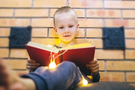 Christmas background. The boy is sitting near the fireplace with a book. Child with yellow lights. Waiting for the new year. Christmas illumination. Brick wall. Bright smileの写真素材