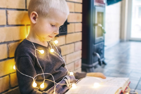 Christmas background. The boy is sitting near the fireplace with a book. Child with yellow lights. Waiting for the new year. Christmas illumination. Brick wall. Bright smileの写真素材