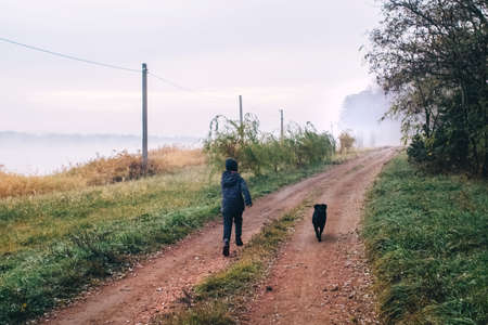 Boy and dog in the forest. A cocker spaniel is running alongside the child along a country road. Walk with a black pet among the autumn trees.の写真素材
