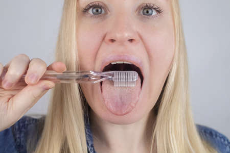 Clean tongue. Close-up of a woman performing antimicrobial hygiene with a special tongue brush. Isolated on white background. A blond female looks into the frame, as if he is looking in a mirror.の写真素材