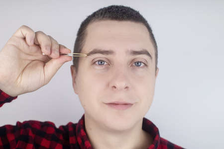 Man plucking eyebrows. Close-up of removing excess hair with tweezers. Isolated on white background. A male looks into the frame and it feels like he is looking in the mirrorの写真素材