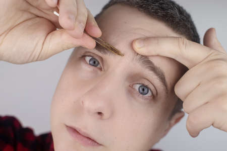 Man plucking eyebrows. Close-up of removing excess hair with tweezers. Isolated on white background. A male looks into the frame and it feels like he is looking in the mirrorの写真素材