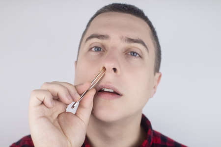 Man pulls out his nose hair with tweezers. Close-up of male hygiene products. Isolated on white background. The male looks at the screen as if in a mirror.の写真素材