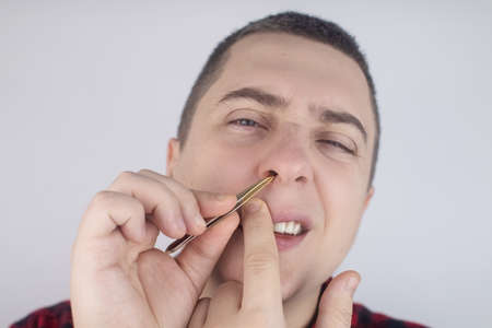 Man pulls out his nose hair with tweezers. Close-up of male hygiene products. Isolated on white background. The male looks at the screen as if in a mirror.の写真素材