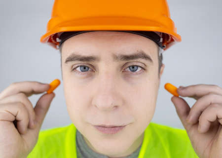 A worker in a hard hat and green vest puts on ear plugs. industrial safety. industrial noise protection. Compliance with the rules of conduct at loud work. Orange ear plugsの写真素材