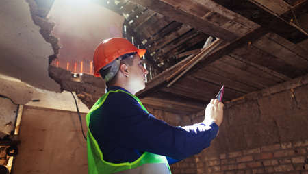 building inspector. Man in a hard hat and a yellow reflective vest examines damaged structures and inspects the building. damage assessment. Preparing for the repair or construction of a buildingの写真素材
