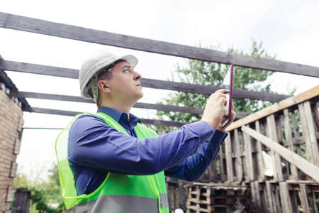 building inspector. Man in a hard hat and a yellow reflective vest examines damaged structures and...