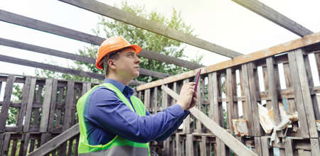 building inspector. Man in a hard hat and a yellow reflective vest examines damaged structures and inspects the building. damage assessment. Preparing for the repair or construction of a buildingの写真素材