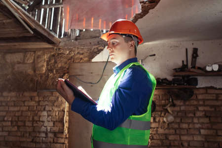building inspector. Man in a hard hat and a yellow reflective vest examines damaged structures and inspects the building. damage assessment. Preparing for the repair or construction of a buildingの写真素材