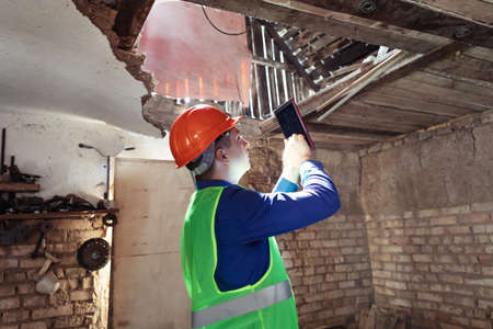 building inspector. Man in a hard hat and a yellow reflective vest examines damaged structures and inspects the building. damage assessment. Preparing for the repair or construction of a buildingの写真素材