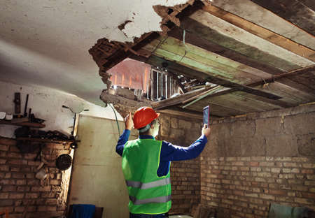 building inspector. Man in a hard hat and a yellow reflective vest examines damaged structures and inspects the building. damage assessment. Preparing for the repair or construction of a buildingの写真素材