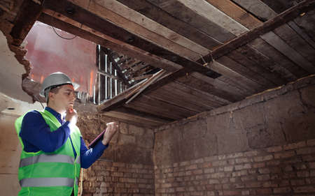 building inspector. Man in a hard hat and a yellow reflective vest examines damaged structures and inspects the building. damage assessment. Preparing for the repair or construction of a buildingの写真素材