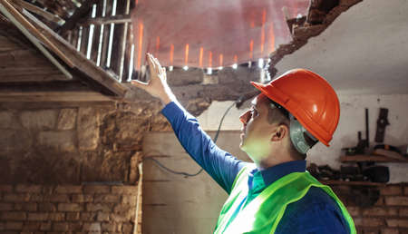 building inspector. Man in a hard hat and a yellow reflective vest examines damaged structures and inspects the building. damage assessment. Preparing for the repair or construction of a buildingの写真素材