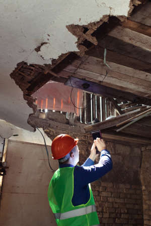 building inspector. Man in a hard hat and a yellow reflective vest examines damaged structures and inspects the building. damage assessment. Preparing for the repair or construction of a buildingの写真素材