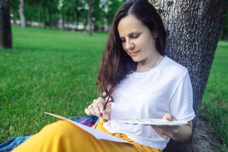 art therapy. Woman draws in the park. Girl sits with her back against a tree and looks into distance. Restoration of nervous system with help of drawing. Concept of relaxation and meditative stateの写真素材