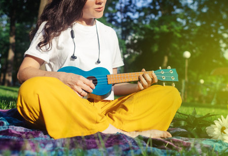 Girl in the park sitting on the grass plays the ukulele. Learning to play a four-string instrument. Relaxation in a public park. Music therapy, psychology, meditation, relaxation, relaxationの写真素材