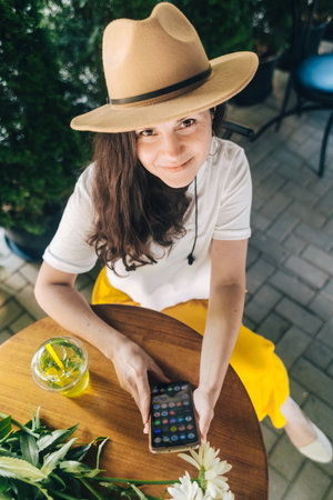 Happy smiling girl sits at a table in a cafe after a romantic date. There is a bouquet of daisies on the table. Woman in a brown hat looks at her phone, reads correspondence and remembers happy momentsの写真素材