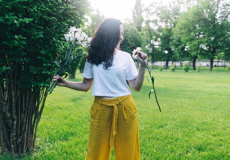 A girl in the park near a large bush sniffs a bouquet of daisies. Good luck, happy pastime, good mood. The concept of healthy relationships, mental health and self-loveの写真素材