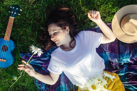 Woman lies on a purple blanket. On her body is a bouquet of daisies. Meditative state in a public park. On the background of the ukulele, a small guitar. Nervous system treatment concept, nature therapyの写真素材