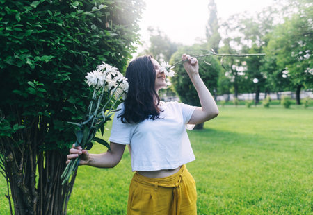 A girl in the park near a large bush sniffs a bouquet of daisies. Good luck, happy pastime, good mood. The concept of healthy relationships, mental health and self-loveの写真素材