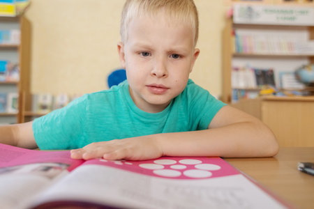 boy in library. Seven year Ukrainian guy is sitting at a table in the library and reading book. Concept of normal learning, thirst for knowledge, desire to learn new things. Blurred background and lettersの写真素材
