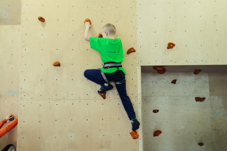 Rock climbing for children. Blond boy in a green shirt is climbing wall. Bouldering class. Artificial track for safe activities and competitions. Mountaineering, safety system, special shoesの写真素材