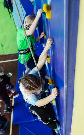 Rock climbing for children. Blond boy in a green shirt is climbing wall. Bouldering class. Artificial track for safe activities and competitions. Mountaineering, safety system, special shoesの写真素材