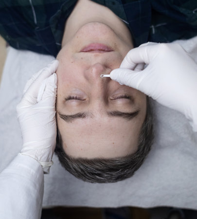 Man undergoing facial cleansing and blackhead removal. Beautician using comedone extractor or uno...