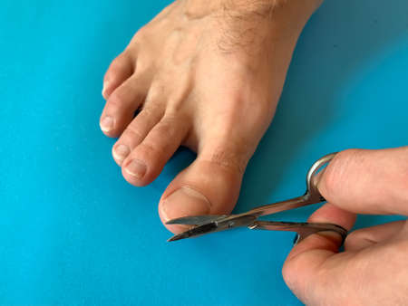 Male's hand holding on nail scissors, Man use nail scissors cutting their thumbs nail on foot toes on blue backgroundの写真素材