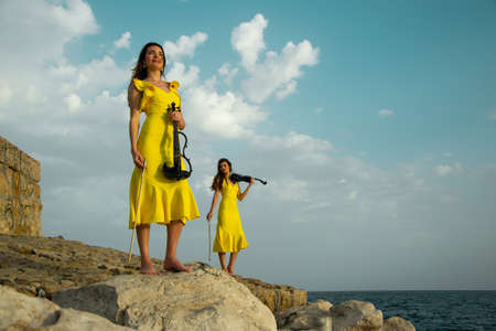 Two beautiful twin sisters violinists in yellow concert dresses are playing electric violins on rocks by the Mediterranean sea in Antalya, Turkey. Sea and cloudy sky on the background. Stock image.の写真素材