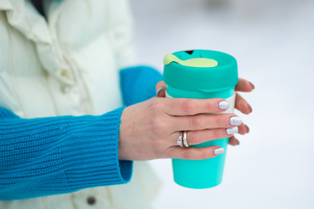 Female's hands holding a green thermal mug in a winter day with a white snow on a background.