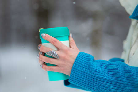 Female's hands holding a green thermal mug in a winter day with a white snow on a background.