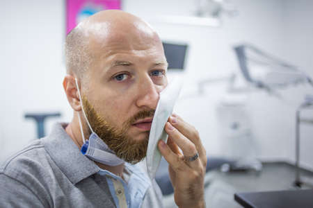 Unhappy and scared caucasian male in a dental clinic waiting for a check up and holding a cooling element at his cheek because of a toothache.の写真素材