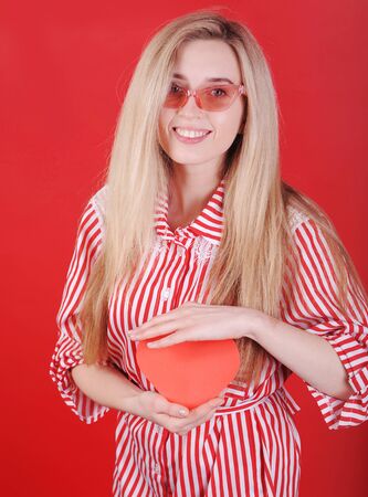 Smiling woman in red dress with paper heart in hand posing on red background. Valentine`s Day concept.の写真素材