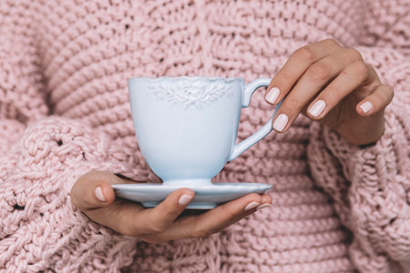 A girl with a fashionable pink manicure holds a blue mug against the background of a knitted pink sweaterの写真素材