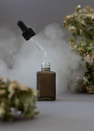 Dropper bottle in white smoke and tree branches on grey background. skin care products.の写真素材