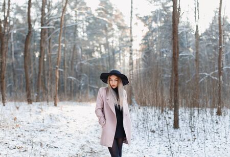 Pretty fashionable woman in black hat and pink coat in winter amazing woods at sunset