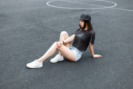 Stylish beautiful woman in black fashionable clothes with a cap sitting on stadium