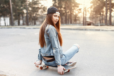 Young beautiful fashionable girl in stylish clothing sitting on a skateboard at sunsetの写真素材