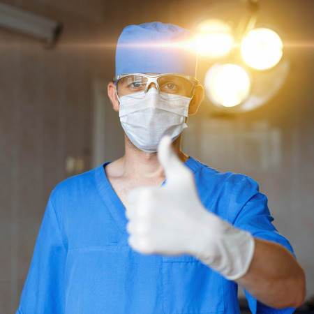 Young specialist surgeon in a mask, glasses and blue uniform holding thumbs up. The concept of a successful doctor.の写真素材