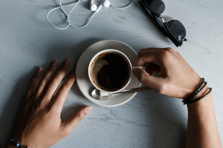 Man drinks coffee. Cup of coffee, headphones and sunglasses lying on a wooden table. Top view . Male hand holding a coffee.の写真素材