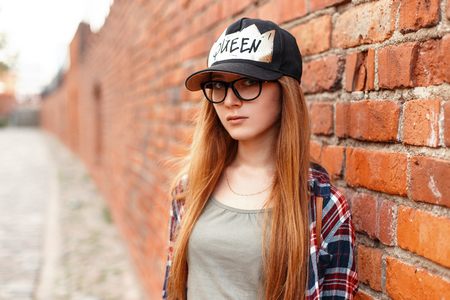 Stylish pretty hipster woman with glasses and a cap near a red brick wall.の写真素材