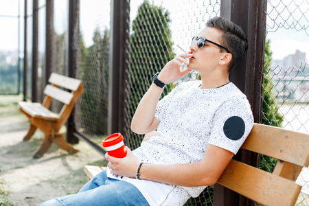 Young depressed man sitting on a bench and smokes. Guy holding a cup of coffee in his hand.の写真素材