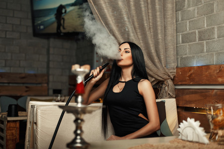 The beautiful girl smokes a hookah and lets the smoke. Woman resting in a barの写真素材