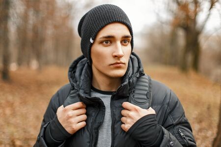 Stylish young man in a black knitted hat and jacket standing near the autumn trees.の写真素材